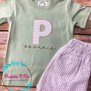 May include: A light green short-sleeved shirt with a white and brown striped letter "P" embroidered on the front. The shirt has the name "phoenix" embroidered below the letter. The shirt is paired with beige and white striped shorts.