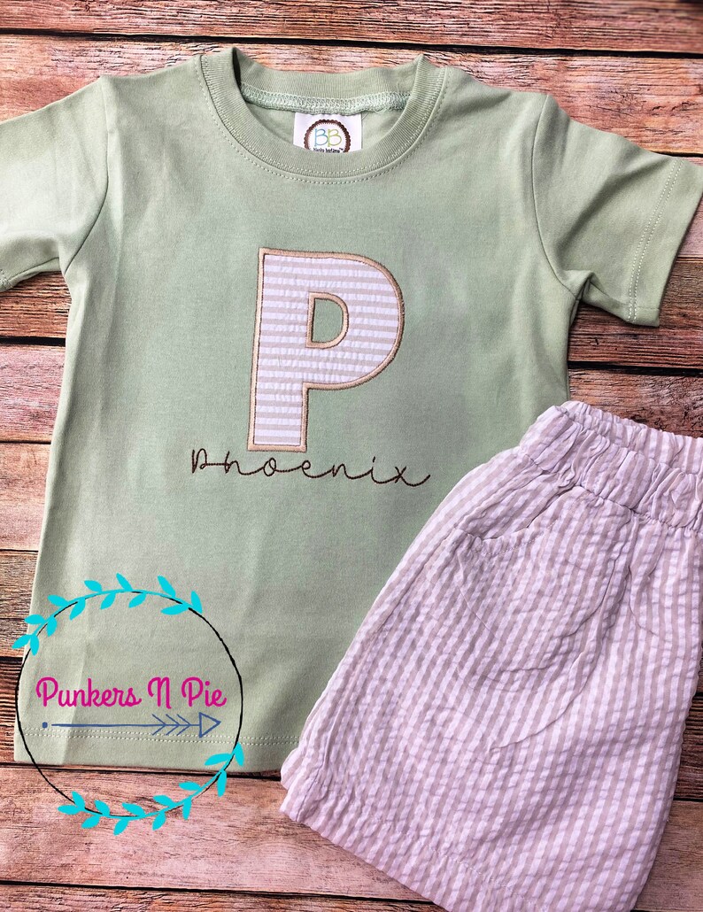 May include: A light green short-sleeved shirt with a white and brown striped letter "P" embroidered on the front. The shirt has the name "phoenix" embroidered below the letter. The shirt is paired with beige and white striped shorts.