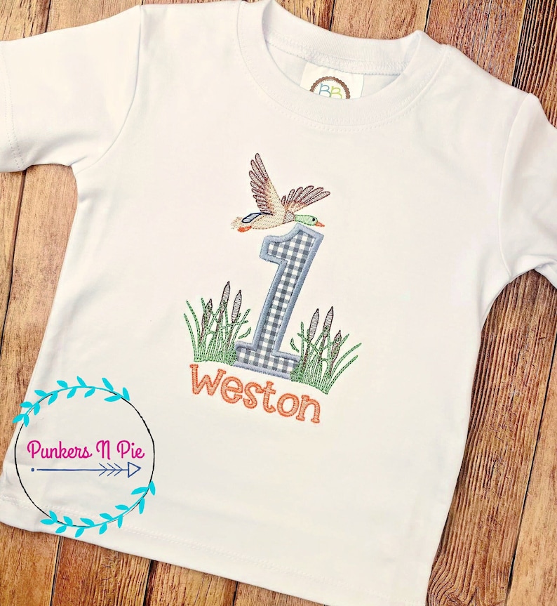 May include: White short-sleeved t-shirt with an embroidered design. The design features a flying duck above a number 1 in a blue and white checkered pattern, with the name "Weston" in orange below.