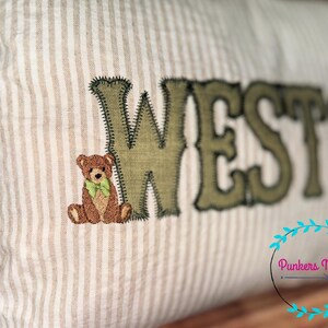 May include: Cream-colored pillow with vertical stripes, featuring the embroidered name "WESTON" in olive green. A small teddy bear with a green bow tie is embroidered next to the letter "W". The pillow has a rustic, handmade aesthetic.