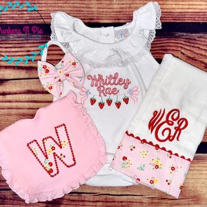 May include: A baby clothing set featuring a white bodysuit with the name "Whitley Rae" and strawberry embroidery, a pink bib with a floral "W", a matching bow, and a white towel with red monogram. The set has a strawberry theme.