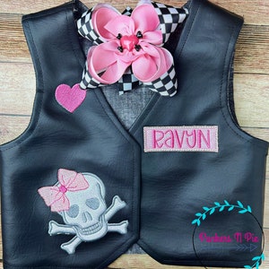 K&ouml;nnte beinhalten: Eine schwarze Kunstlederweste mit einem rosa Herzen, einem Totenkopf mit gekreuzten Knochen mit einer rosa Schleife und einem rosa Namensschild mit der Aufschrift "RAVYNN".
