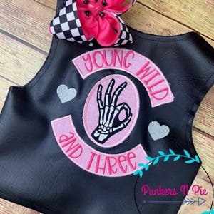 May include: Black leather vest with pink embroidered patches that say "Young Wild and Three" with a skeleton hand making the OK sign in the center.