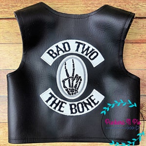 K&ouml;nnte beinhalten: Schwarze Lederweste mit wei&szlig;en gestickten Patches, auf denen "Bad Two The Bone" steht, und einer Skeletthand, die ein Peace-Zeichen macht.