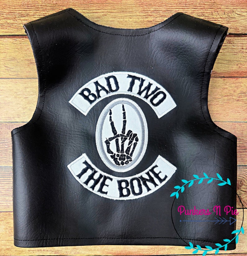 K&ouml;nnte beinhalten: Schwarze Lederweste mit wei&szlig;en gestickten Patches, auf denen "Bad Two The Bone" steht, und einer Skeletthand, die ein Peace-Zeichen macht.