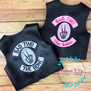 May include: Two black leather vests with embroidered patches. The patches are white and pink with the text "Bad Two the Bone" and a skeleton hand making a peace sign.
