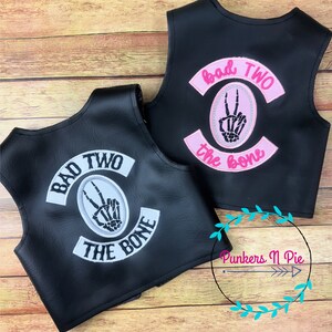 K&ouml;nnte beinhalten: Zwei schwarze Lederwesten mit gestickten Patches. Die Patches sind wei&szlig; und rosa mit dem Text "Bad Two the Bone" und einer Skeletthand, die ein Peace-Zeichen macht.