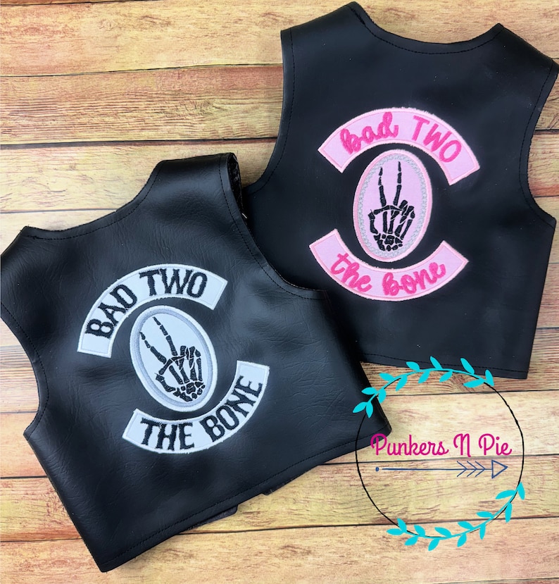 K&ouml;nnte beinhalten: Zwei schwarze Lederwesten mit gestickten Patches. Die Patches sind wei&szlig; und rosa mit dem Text "Bad Two the Bone" und einer Skeletthand, die ein Peace-Zeichen macht.