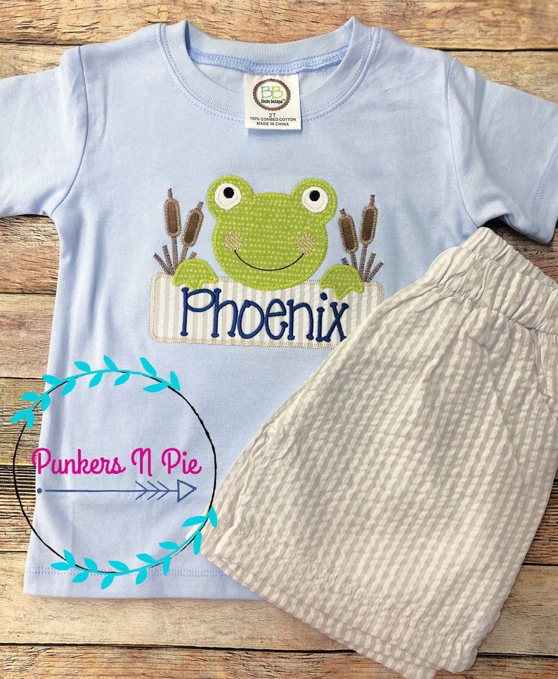 May include: A light blue short-sleeved t-shirt with a green frog applique and the name "Phoenix" embroidered on a brown banner. The shirt is paired with beige striped shorts.