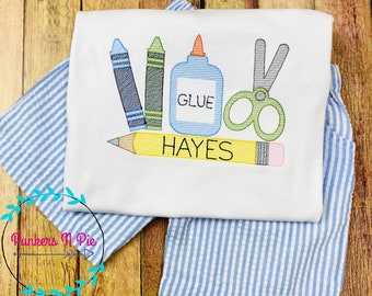 May include: White t-shirt with embroidered school supplies: blue and green crayons, a glue bottle with "GLUE" text, and green scissors. A yellow pencil with "HAYES" text is at the bottom. The shirt is on blue and white striped fabric.