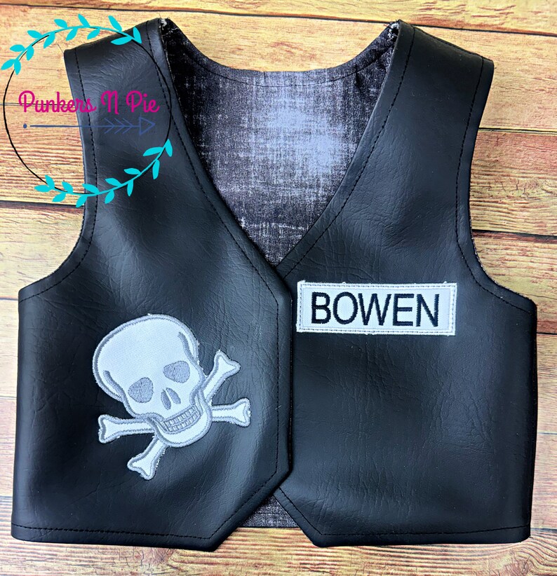K&ouml;nnte beinhalten: Schwarze Kunstlederweste mit einem wei&szlig;en Totenkopf- und Knochenkreuz-Design auf der linken Seite und einem wei&szlig;en Patch mit dem Namen "BOWEN" auf der rechten Seite.