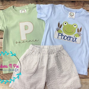 May include: A light blue and green t-shirt with a frog and the name "Phoenix" embroidered on it. A light green t-shirt with a white and brown letter "P" and the name "Phoenix" embroidered on it. A pair of beige shorts with white stripes.