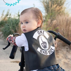 May include: A baby wearing a black leather vest with a white patch that says "ONE BAD BOY" and a skeleton hand making a peace sign. The baby is sitting on a black motorcycle.