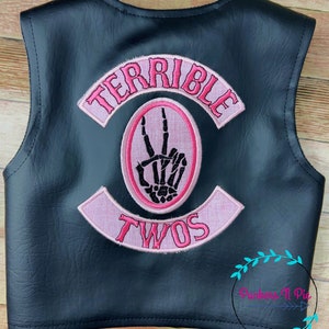 May include: Black leather vest with pink embroidered patches that say "Terrible Twos" and feature a skeleton hand making a peace sign.