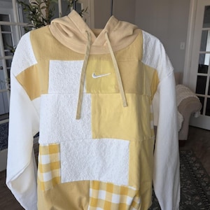 May include: A patchwork hoodie in white and yellow, featuring a small Nike logo. The design incorporates various textures, including a gingham pattern and a textured white fabric. The hood and sleeves are solid colors.