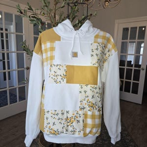 May include: A white hooded sweatshirt featuring a patchwork design of solid mustard yellow, yellow and white gingham check, and a yellow floral print on a white background. The hoodie includes a small rectangular Carhartt logo patch on the chest.