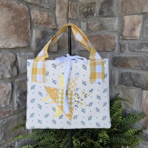 May include: A square fabric tote bag featuring a cream background with a small blue floral print. The center displays a patchwork star in yellow and white gingham and floral patterns. It has yellow and white gingham handles and a white lace bow.