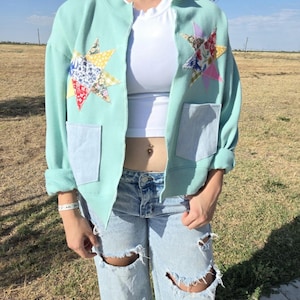 May include: A light mint green bomber jacket featuring two large star-shaped patches made from assorted floral and patterned fabric scraps on the chest, along with two light blue rectangular pockets on the lower front panels and ribbed trim.