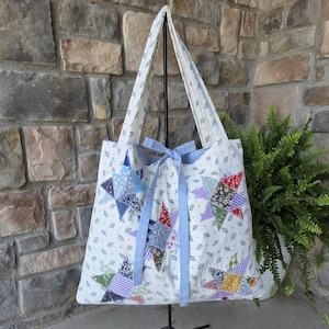 May include: A white fabric tote bag featuring a small blue floral print and multiple patchwork star designs in various colors and patterns. The bag includes two long shoulder straps and a light blue ribbon tie closure at the top center.