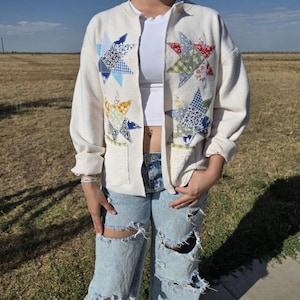May include: An off-white long-sleeve cardigan featuring four large star-shaped patchwork appliqués in various blue, yellow, red, and green floral and gingham patterns. The garment includes two front pockets and a simple open-front design.