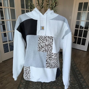 May include: A long-sleeve hooded sweatshirt featuring a patchwork design of white, grey, and black fabric panels. The front includes sections with a brown and white zebra print pattern and a small rectangular Carhartt logo patch at the center chest.