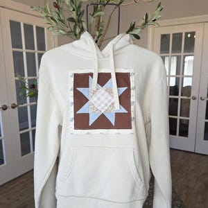 May include: A cream-colored hooded sweatshirt featuring a central square patch with a brown background and a multi-pointed star design. The star is composed of light blue, beige gingham, and floral patterned fabric segments. It has a front pocket.