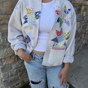 May include: A beige open-front sweatshirt featuring long sleeves and two front pockets. The garment is decorated with large, multi-colored patchwork star appliqués in various floral, striped, and checkered patterns. The pockets have a red and white trim.