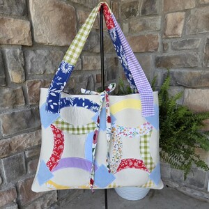 May include: A tote bag featuring a cream-colored base with a multi-colored double wedding ring quilt pattern. The bag includes two shoulder straps with green gingham and floral prints, and a central tie closure with a floral fabric pattern.