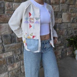 May include: A cream-colored long-sleeve cardigan featuring colorful fabric star appliques in various patterns and a light blue denim patch pocket on the front. The cardigan is worn over a plain white crew-neck t-shirt and light blue denim jeans.