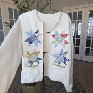 May include: An off-white long-sleeve cardigan featuring four appliquéd star motifs made from various patterned fabrics in shades of blue, red, green, and yellow. The garment includes two quilted off-white patch pockets with green gingham trim.