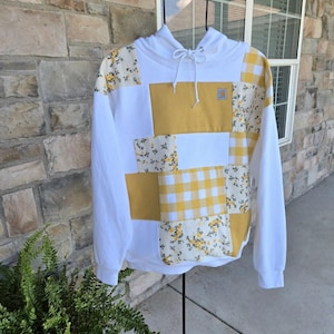 May include: A white hooded sweatshirt featuring a patchwork design on the front. The pattern includes solid mustard yellow panels, yellow and white gingham checks, and a white fabric with a small yellow floral print. A small Carhartt logo is visible.