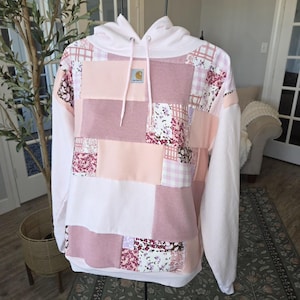 May include: A patchwork hoodie in shades of pink, white, and floral patterns. The hoodie features a white hood with drawstrings, white sleeves, and a Carhartt logo patch. The patchwork design includes squares of solid pink, floral, and gingham fabrics.