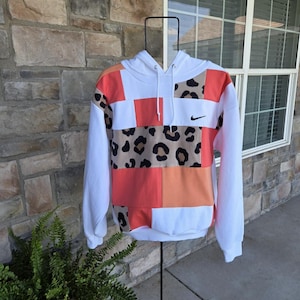 May include: A long-sleeve hooded sweatshirt featuring a patchwork design of white, coral, and leopard print fabric panels. The garment includes a drawstring hood, ribbed cuffs, and a small black Nike swoosh logo embroidered on the upper chest area.