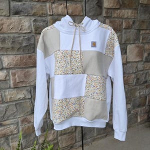 May include: A patchwork hoodie featuring a mix of solid white, beige, and small floral print fabric panels. The garment includes a hood with beige drawstrings and a small rectangular Carhartt brand patch on the upper left chest area.