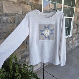 May include: A light beige heathered long-sleeve sweatshirt featuring a centered square fabric appliqué. The appliqué displays a blue star pattern with a central white square containing a small yellow and green floral print on a tan checkered background.