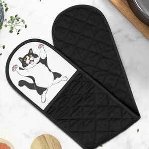 May include: Black and white cat oven mitt with a quilted black back. The cat is depicted with its arms raised in the air, as if it is surprised.