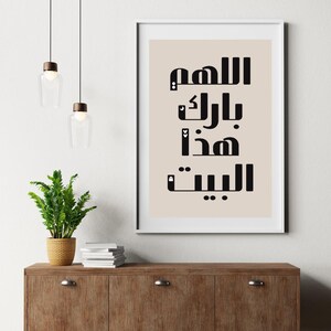 Op de afbeelding: Een ingelijste kunstdruk met zwarte Arabische kalligrafie op een beige achtergrond. De print wordt getoond op een houten kast, met een kleine potplant en hanglampen op de achtergrond. De tekst luidt "O Allah, zegen dit huis."
