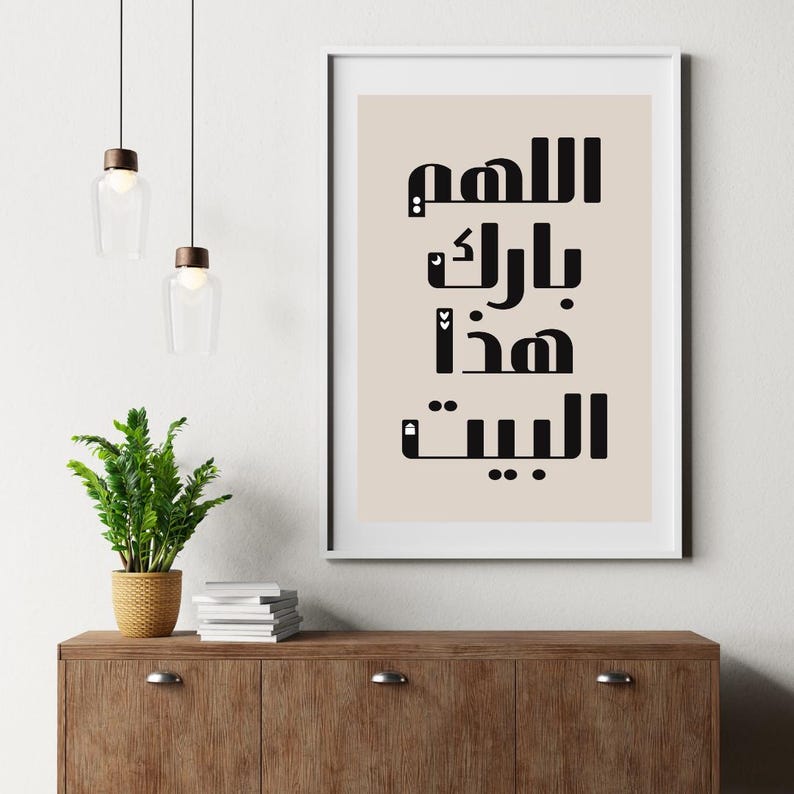 Op de afbeelding: Een ingelijste kunstdruk met zwarte Arabische kalligrafie op een beige achtergrond. De print wordt getoond op een houten kast, met een kleine potplant en hanglampen op de achtergrond. De tekst luidt "O Allah, zegen dit huis."