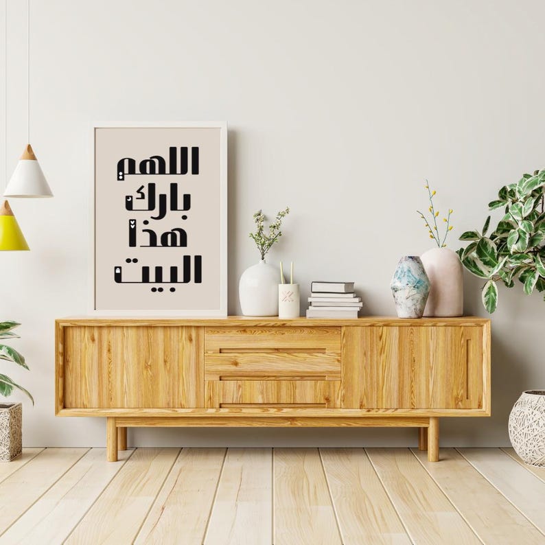 Op de afbeelding: Een ingelijste print met zwarte Arabische kalligrafie op een beige achtergrond, geplaatst op een licht houten kast. De kast heeft lades en deuren, met vazen, boeken en planten erop. De kamer heeft een minimalistisch ontwerp.