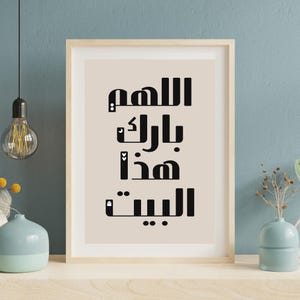 Op de afbeelding: Ingelijste kunstafdruk met zwarte Arabische kalligrafie op een beige achtergrond. De tekst in het midden luidt: "Allah, zegen dit huis". De lijst is van licht hout en de print wordt op een witte plank gepresenteerd.