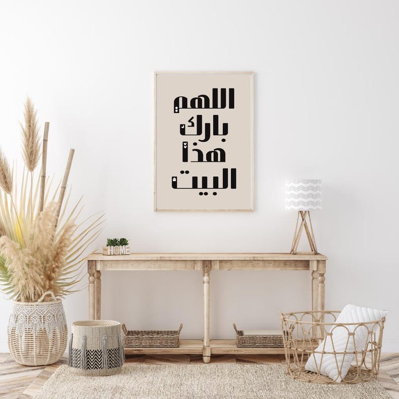 Op de afbeelding: Een ingelijste print met zwarte Arabische kalligrafie op een beige achtergrond. De print hangt boven een houten console met decoratieve manden en een lamp. De kamer is ingericht in neutrale tinten met natuurlijke texturen.