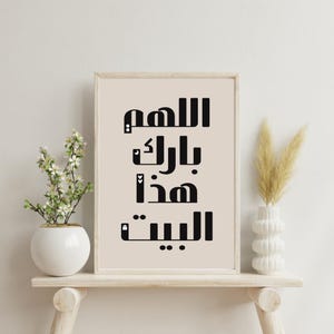 Op de afbeelding: Een ingelijste print met zwarte Arabische kalligrafie op een beige achtergrond. De verticaal geplaatste tekst luidt "Allah, zegen dit huis". De print staat op een licht houten plank, met een witte vaas met bloemen links en een witte vaas met gedroogd gras rechts.