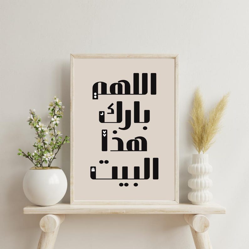 Op de afbeelding: Een ingelijste print met zwarte Arabische kalligrafie op een beige achtergrond. De verticaal geplaatste tekst luidt "Allah, zegen dit huis". De print staat op een licht houten plank, met een witte vaas met bloemen links en een witte vaas met gedroogd gras rechts.