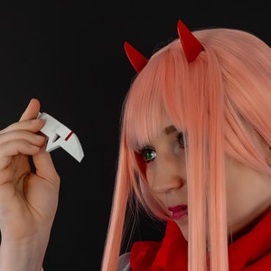 Könnte beinhalten: Eine Frau mit langen, rosafarbenen Haaren und roten Teufelshörnern trägt einen roten Schal und eine graue Jacke. Sie hält ein weißes Objekt in der Hand.