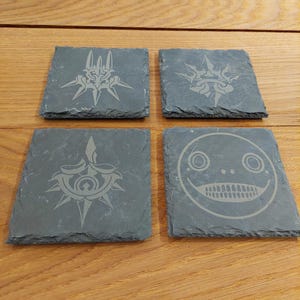 May include: Four black slate coasters with different designs. The coasters feature a stylized design of a sword, a face, and two different versions of a crown.