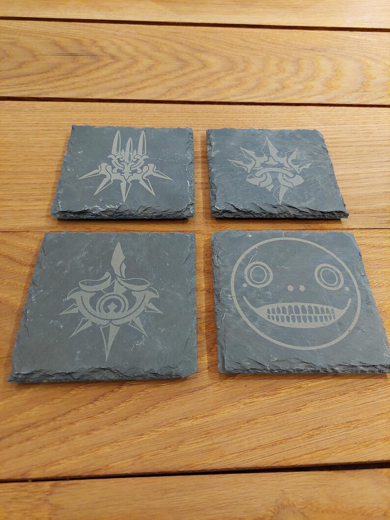 May include: Four black slate coasters with different designs. The coasters feature a stylized design of a sword, a face, and two different versions of a crown.