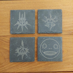 May include: Set of four black slate coasters with etched designs. The designs include a crown with spikes, a face with large eyes and a wide mouth, and two different versions of a star with spikes.