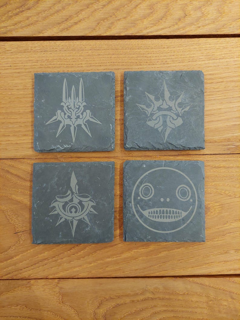 May include: Set of four black slate coasters with etched designs. The designs include a crown with spikes, a face with large eyes and a wide mouth, and two different versions of a star with spikes.