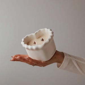 Puede incluir: Una vela blanca en forma de corazón de cerámica con dos mechas. La vela está siendo sostenida en una mano.
