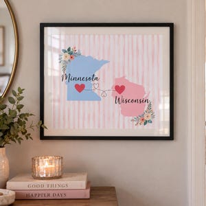 Op de afbeelding: Een ingelijste kunstprint met een lichtblauw silhouet van Minnesota en een roze silhouet van Wisconsin op een verticaal roze gestreepte achtergrond. De staten zijn verbonden door een stippellijn met twee rode harten en bloemaccenten.